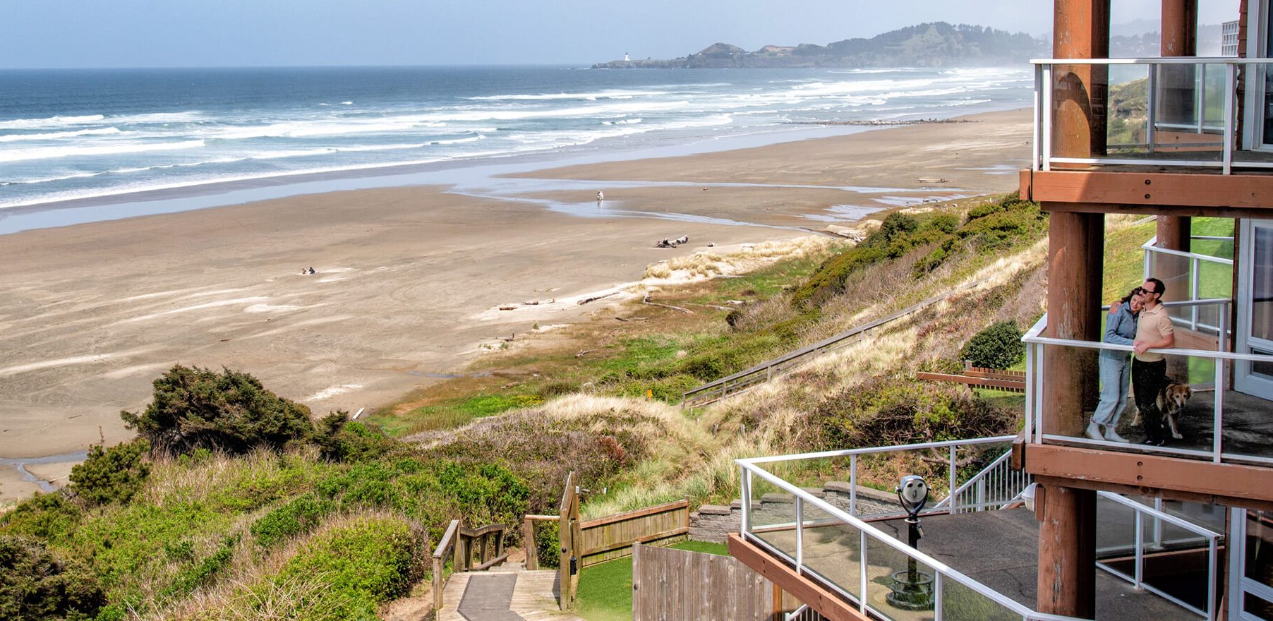 The beach and ocean meeting with a hotel to the right and people hugging on a balcony of one of the rooms