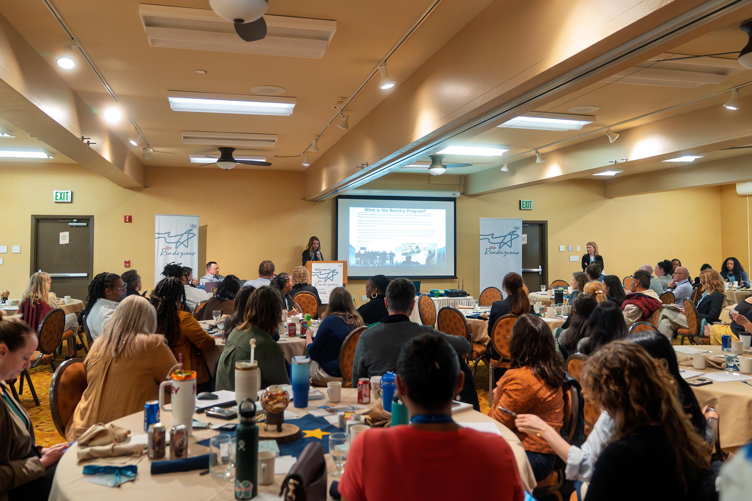 Conference room full of people with a speaker giving a presentation at the front